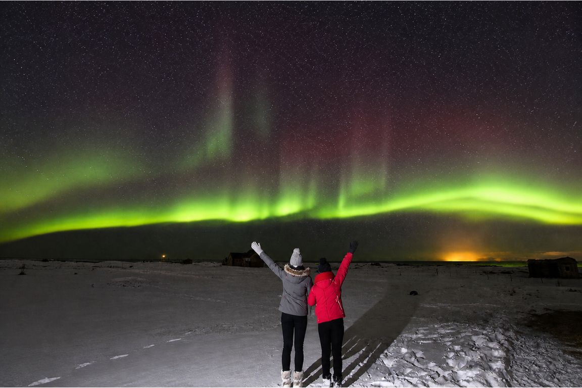 Northern Lights dancing over snowy Iceland landscape with two visitors enjoying the view during a Day Trips From Reykjavik tour with our agency