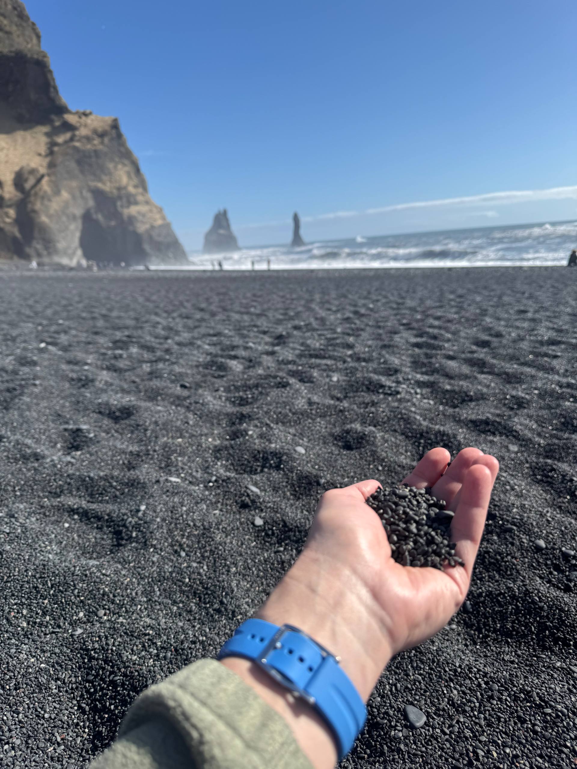 Reynisfjara Black Sand Beach in Iceland with volcanic pebbles in hand and Reynisdrangar sea stacks in the distance during a Day Trips From Reykjavik guided tour with our agency