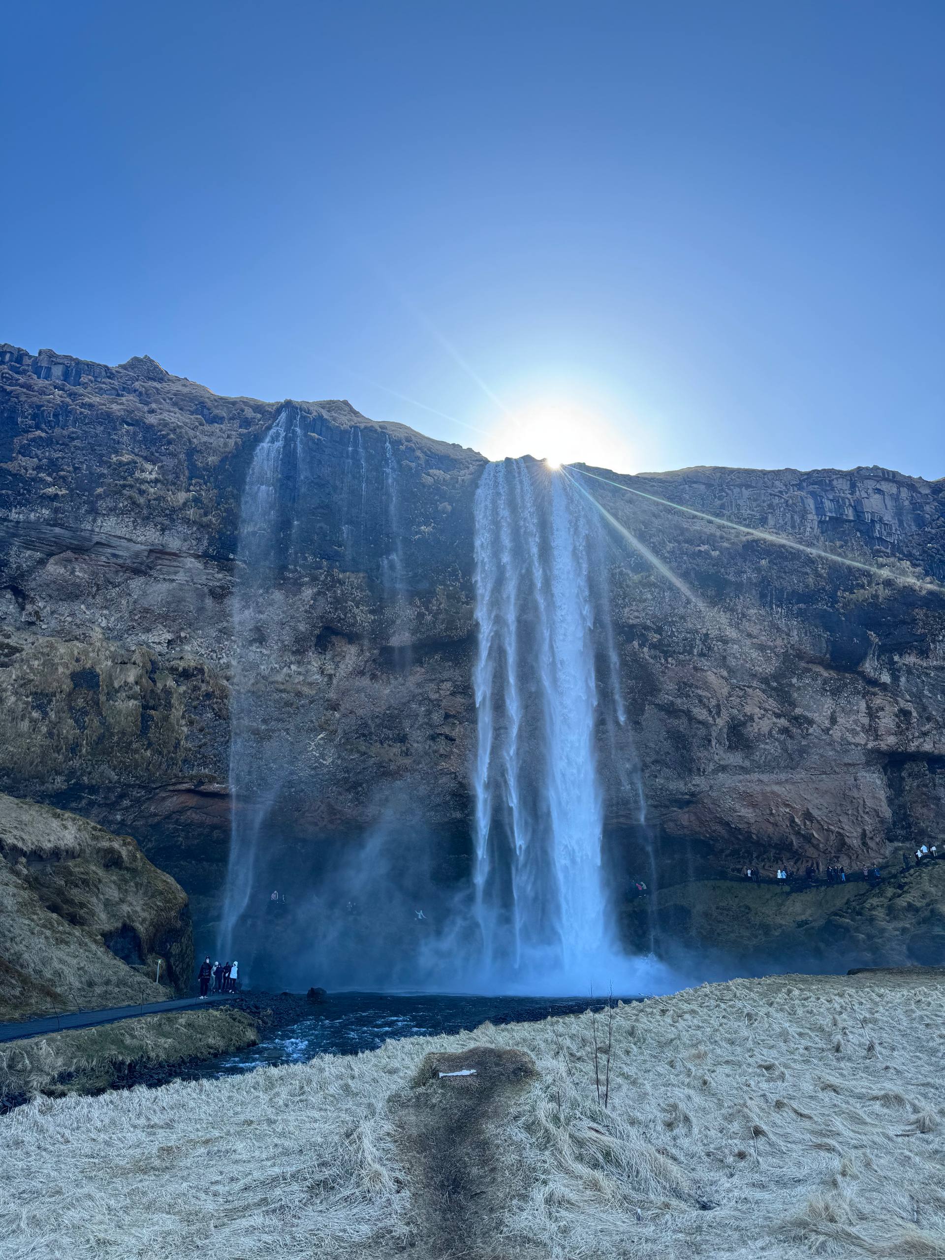 Seljalandsfoss waterfall flowing into a river with scenic cliff backdrop and visitors exploring the area during a Day Trips From Reykjavik tour with our agency
