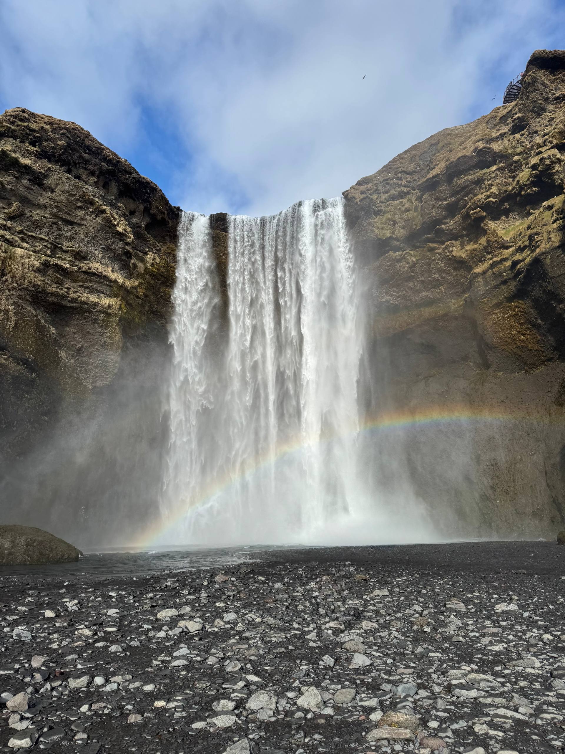 Skógafoss waterfall in Iceland with a vivid rainbow at its base and rocky foreground during a Day Trips From Reykjavik tour with our agency