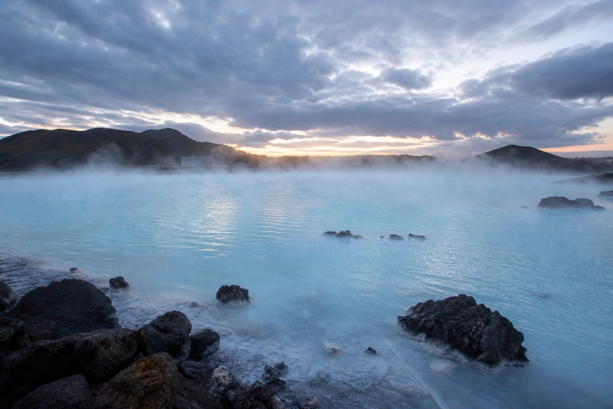 Steam rising off a geothermal lagoon at dusk with a dark lava shoreline